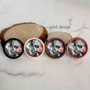 May include: Four round, wooden-framed pins featuring a grayscale image of a scary clown face with red accents. The frames are black, natural wood, dark brown, and orange. The text "---johill design" is visible.