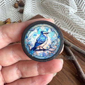 Op de afbeelding: Ronde houten speld met een glas-in-lood ontwerp van een blauwe gaai op een tak met blauwe bloemen. De vogel heeft blauwe en witte veren. De speld heeft een donkere rand en de tekst "johill design".