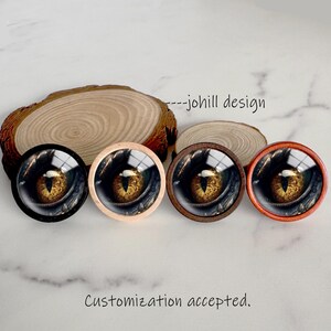 Könnte beinhalten: Vier runde dekorative Schrankknöpfe mit einem detaillierten Drachenaugen-Design. Die Knöpfe haben schwarze, natürliche Holz-, dunkelbraune und orangefarbene Rahmen. Der Text "---johill design" und "Customization accepted" sind sichtbar.