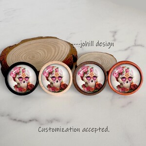 May include: Four decorative skull designs with floral and bow accents. Each skull features a unique design with pink and red roses. The skulls are set in round frames with black, light wood, dark wood, and orange finishes. The text "--johill design" is visible.