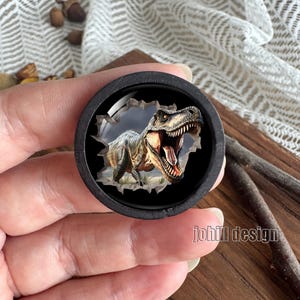 Op de afbeelding: Een rond, zwart houten object met een 3D-afbeelding van een Tyrannosaurus Rex die door een gescheurd oppervlak breekt. De dinosaurus heeft een open mond, met tanden en een rood interieur. De tekst "johill design" is zichtbaar.