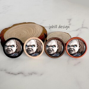 May include: Four round Chucky-themed pinback buttons with different colored frames: black, natural wood, dark brown, and orange. Each button features a close-up of Chucky's face with blue eyes. The background includes a wooden slice and the text "--johill design".