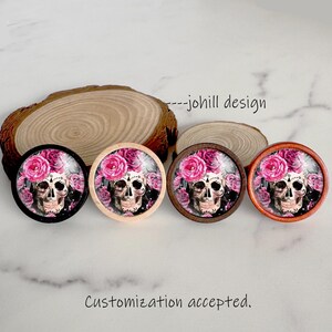May include: Four round decorative skull designs with pink roses. The skulls have intricate details and are surrounded by a floral arrangement. The designs are set in wooden frames with varying colours, including black, light wood, and brown.