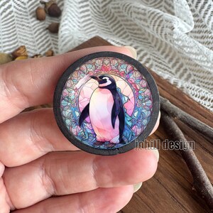 Pode incluir: Uma base de copo redonda de madeira preta com uma ilustração de pinguim. O pinguim tem cabeça e asas pretas, peito branco e barriga rosa, sobre um fundo em estilo vitral. A base de copo tem o texto "johill design".