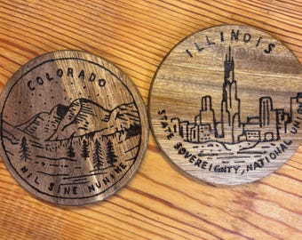 May include: Two round wooden coasters. One coaster features a mountain and forest scene with the word "COLORADO" and the phrase "NIL SINE NUMINE". The other coaster depicts a city skyline with the word "ILLINOIS" and the phrase "STATE SOVEREIGNTY, NATIONAL UNION".