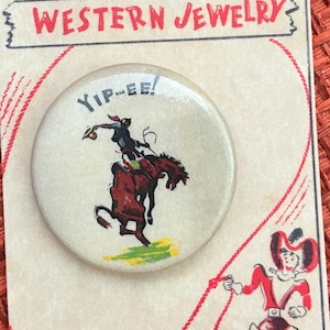 May include: A round, cream-colored button with a cowboy riding a bucking bronco, the words "Yip-ee!" above the image. The button is on a card that reads "Western Jewelry" and "futuravintage.com". The card has a red and white illustration of a cowboy.