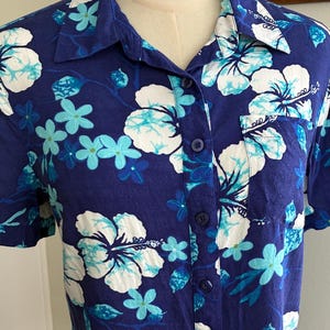 May include: A short-sleeved button-down shirt in a deep blue hue, adorned with a vibrant floral pattern. The design features large white hibiscus flowers with turquoise accents, complemented by smaller turquoise blossoms. The shirt has a classic collar and a single chest pocket.