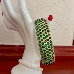 May include: A gold-toned bangle bracelet featuring three rows of circular, vibrant green rhinestones set into the metal surface. The bracelet is displayed on a white, hand-shaped stand that is adorned with a single strand of small white pearls.