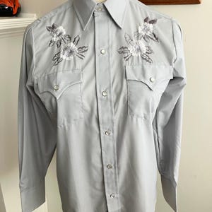May include: A light grey long-sleeved button-down shirt with white embroidered flowers on the chest.