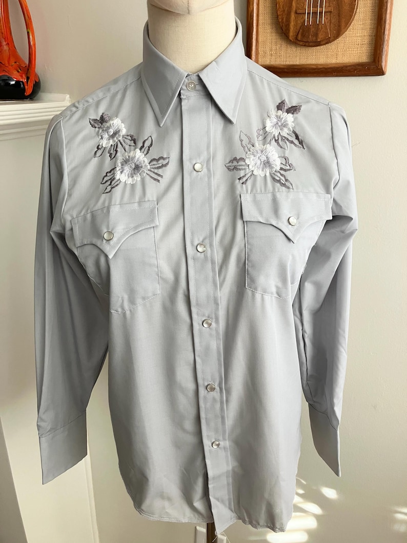 May include: A light grey long-sleeved button-down shirt with white embroidered flowers on the chest.