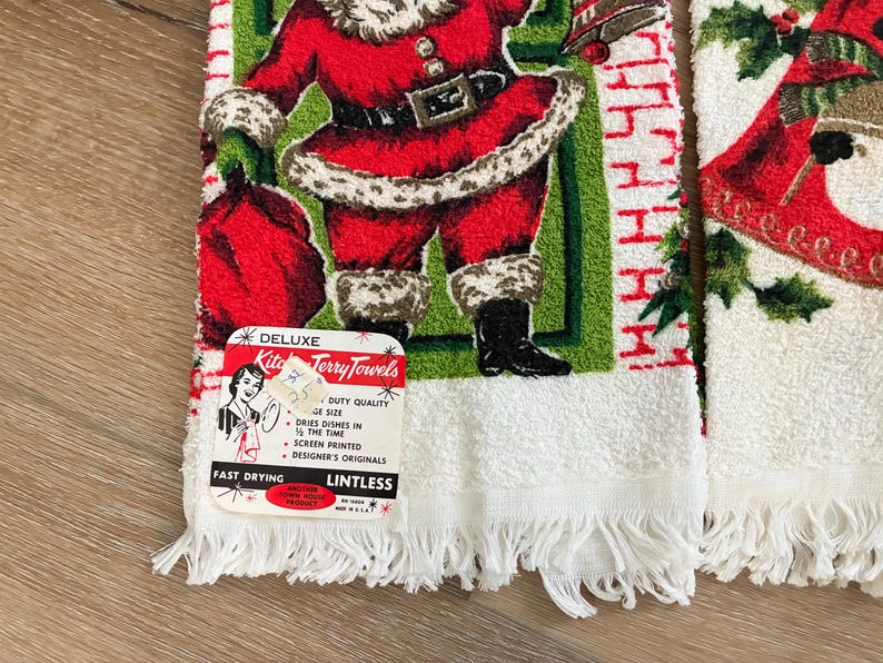K&ouml;nnte beinhalten: Vintage Weihnachts-K&uuml;chent&uuml;cher mit einem Weihnachtsmann-Design. Die Handt&uuml;cher sind wei&szlig; mit einem Fransenrand und zeigen eine rot-gr&uuml;ne Weihnachtsmann-Illustration. Die Handt&uuml;cher haben ein Etikett mit der Aufschrift "Deluxe Kitchen Terry Towels".