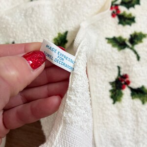 K&ouml;nnte beinhalten: Wei&szlig;es Handtuch mit einem Stechpalmen- und Beerenmuster. Das Handtuch hat einen Fransenbesatz am unteren Rand. Ein Etikett lautet "MADE EXPRESSLY FOR TOWEL DECORATORS".