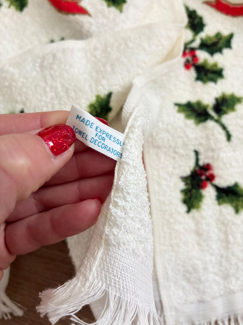K&ouml;nnte beinhalten: Wei&szlig;es Handtuch mit einem Stechpalmen- und Beerenmuster. Das Handtuch hat einen Fransenbesatz am unteren Rand. Ein Etikett lautet "MADE EXPRESSLY FOR TOWEL DECORATORS".