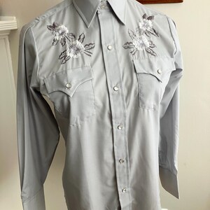 May include: A light grey long-sleeved button-down shirt with embroidered white flowers on the chest.