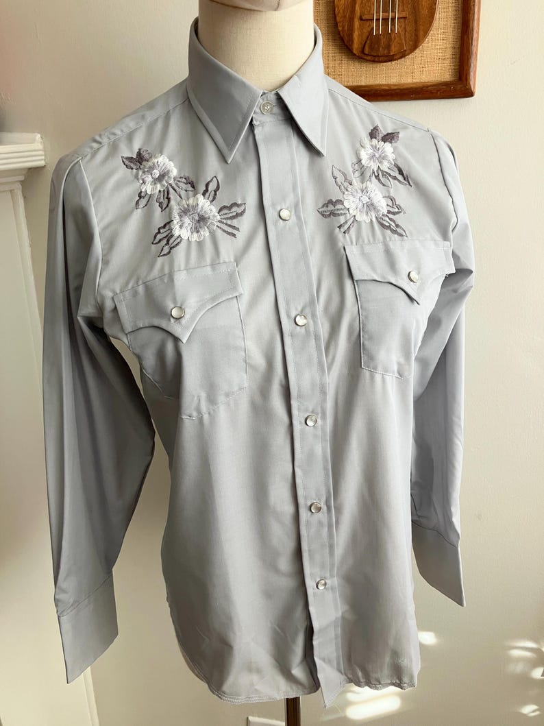 May include: A light grey long-sleeved button-down shirt with embroidered white flowers on the chest.