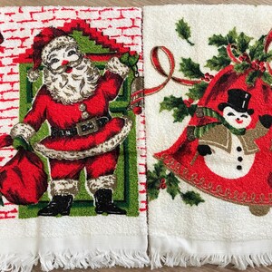 K&ouml;nnte beinhalten: Zwei wei&szlig;e Weihnachtshandtt&uuml;cher. Eines zeigt den Weihnachtsmann in einem roten Anzug mit einem Sack, das andere einen Schneemann in einer Glocke. Beide haben Fransen und festliche Designs.