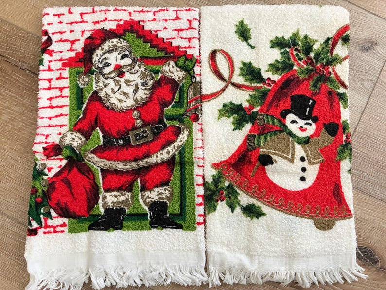 K&ouml;nnte beinhalten: Zwei wei&szlig;e Weihnachtshandtt&uuml;cher. Eines zeigt den Weihnachtsmann in einem roten Anzug mit einem Sack, das andere einen Schneemann in einer Glocke. Beide haben Fransen und festliche Designs.