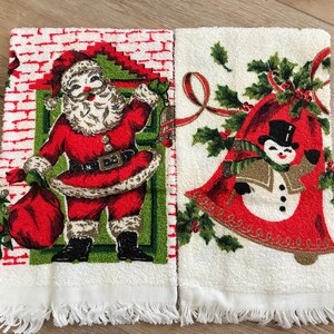K&ouml;nnte beinhalten: Zwei wei&szlig;e Weihnachtshandtt&uuml;cher mit Fransen. Ein Handtuch zeigt den Weihnachtsmann in einem roten Anzug mit einem Sack. Das andere Handtuch hat einen Schneemann in einer roten Glocke mit Stechpalmen und Schleife.