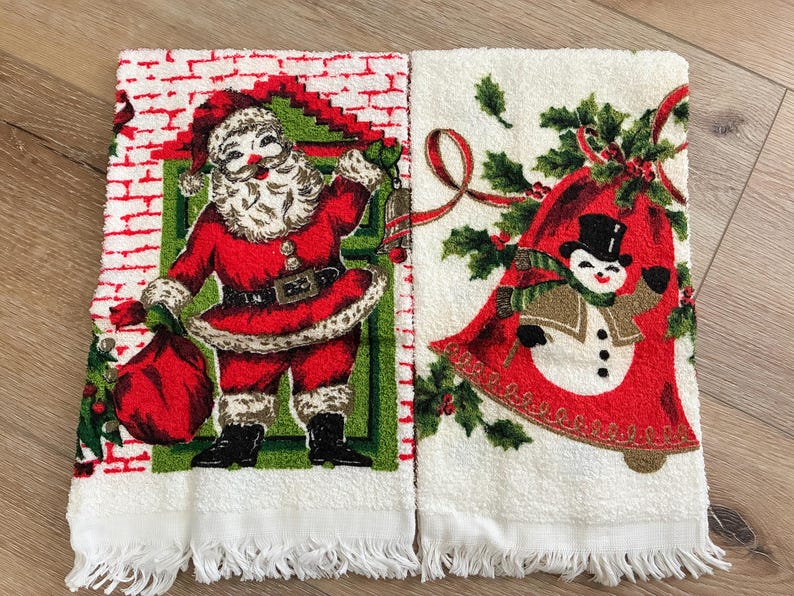 K&ouml;nnte beinhalten: Zwei wei&szlig;e Weihnachtshandtt&uuml;cher mit Fransen. Ein Handtuch zeigt den Weihnachtsmann in einem roten Anzug mit einem Sack. Das andere Handtuch hat einen Schneemann in einer roten Glocke mit Stechpalmen und Schleife.