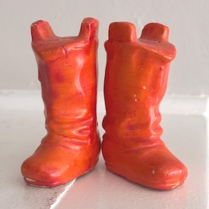 May include: A pair of ceramic cowboy boot figurines in shades of orange. The boots have a classic western design with a rounded toe and a tall shaft. The figurines are likely decorative items, adding a touch of rustic charm.
