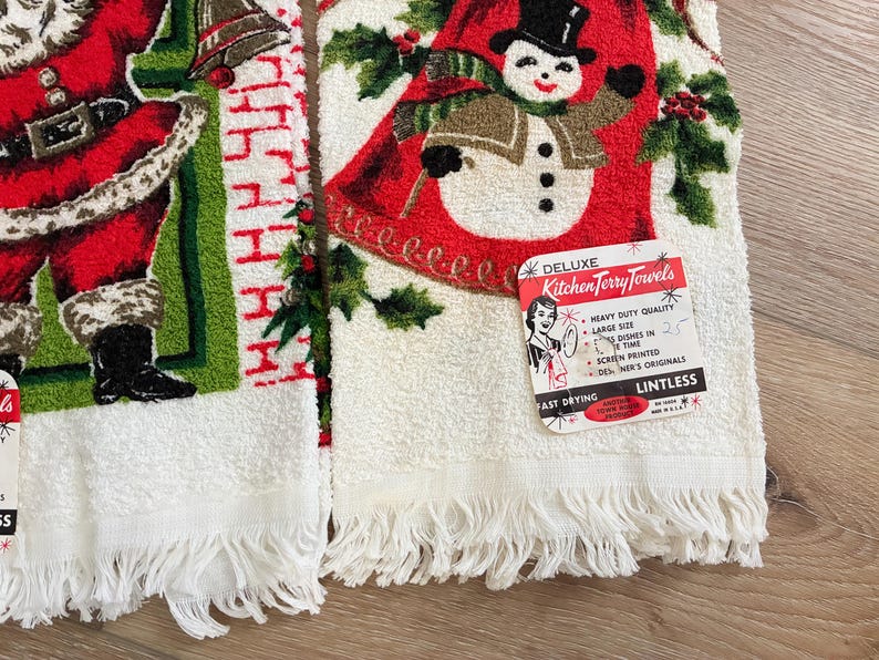 K&ouml;nnte beinhalten: Zwei Vintage-Weihnachts-K&uuml;chent&uuml;cher. Eines zeigt den Weihnachtsmann in einem roten Anzug, das andere einen Schneemann mit Zylinder. Beide haben wei&szlig;e Fransen und den Text "DELUXE Kitchen Terry Towels".