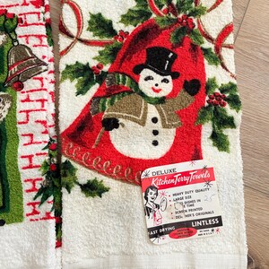 K&ouml;nnte beinhalten: Wei&szlig;es K&uuml;chentuch mit Schneemann-Design, Stechpalme und roten B&auml;ndern. Das Handtuch hat einen Fransenrand und ein Etikett mit der Aufschrift "Deluxe Kitchen Terry Towels". Der Schneemann tr&auml;gt einen schwarzen Zylinder und einen gr&uuml;nen Schal.