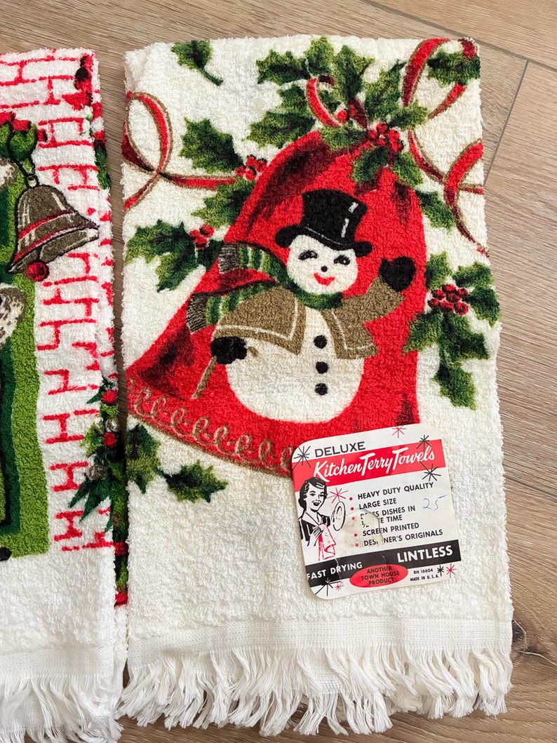 K&ouml;nnte beinhalten: Wei&szlig;es K&uuml;chentuch mit Schneemann-Design, Stechpalme und roten B&auml;ndern. Das Handtuch hat einen Fransenrand und ein Etikett mit der Aufschrift "Deluxe Kitchen Terry Towels". Der Schneemann tr&auml;gt einen schwarzen Zylinder und einen gr&uuml;nen Schal.