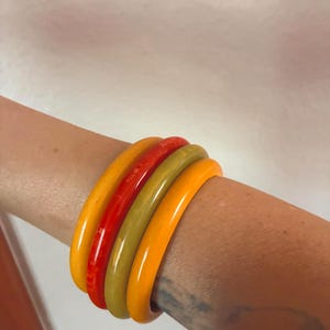 May include: A set of four smooth, rounded bangle bracelets worn together. The stack includes one mustard yellow, one bright red, one olive green, and one vibrant orange bangle, all featuring a glossy, solid-colored finish and a simple circular shape.