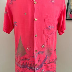 May include: A short-sleeved, coral-colored shirt with a button-down front and a chest pocket. The shirt features a repeating pattern of boats, suns, and birds in shades of purple and brown. The buttons are gold-colored.