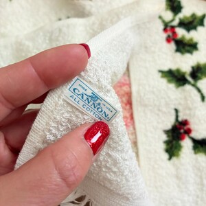 K&ouml;nnte beinhalten: Wei&szlig;es Baumwolltuch mit einem festlichen Stechpalmen- und Bandmuster. Das Handtuch zeigt gr&uuml;ne Stechpalmenbl&auml;tter und rote Beeren mit roten Bandakzenten. Das Etikett lautet "MADE IN USA CANNON ALL COTTON".