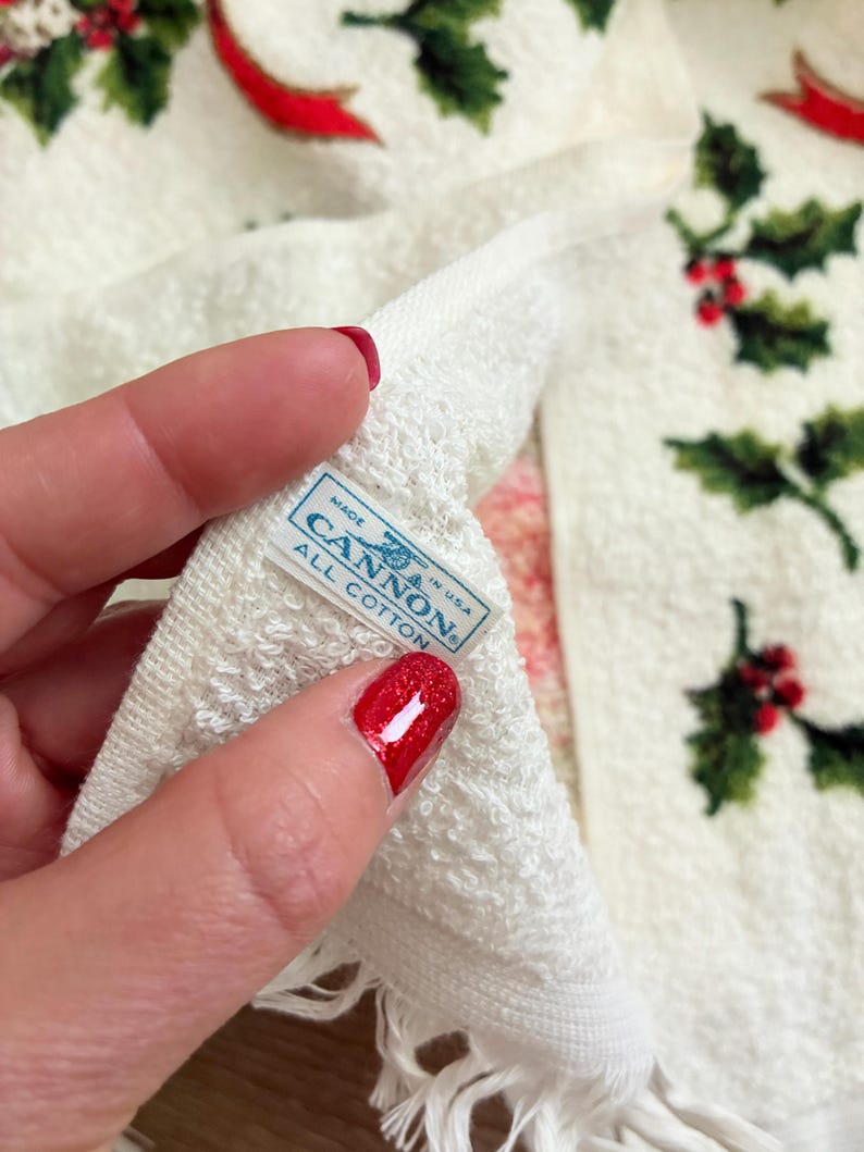 K&ouml;nnte beinhalten: Wei&szlig;es Baumwolltuch mit einem festlichen Stechpalmen- und Bandmuster. Das Handtuch zeigt gr&uuml;ne Stechpalmenbl&auml;tter und rote Beeren mit roten Bandakzenten. Das Etikett lautet "MADE IN USA CANNON ALL COTTON".