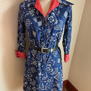 May include: A long-sleeved button-down dress featuring a blue and white bandana-style geometric print. The dress has a red and white polka dot lining visible on the collar and cuffs. A black leather belt with a silver buckle is worn at the waist.