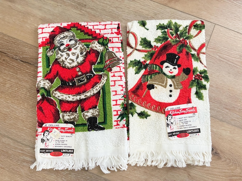K&ouml;nnte beinhalten: Zwei Vintage-Weihnachts-K&uuml;chent&uuml;cher. Eines zeigt den Weihnachtsmann mit Glocke und Sack, das andere einen Schneemann mit Stechpalme. Beide haben wei&szlig;e Fransen und Etiketten mit der Aufschrift "Deluxe Kitchen Terry Towels".