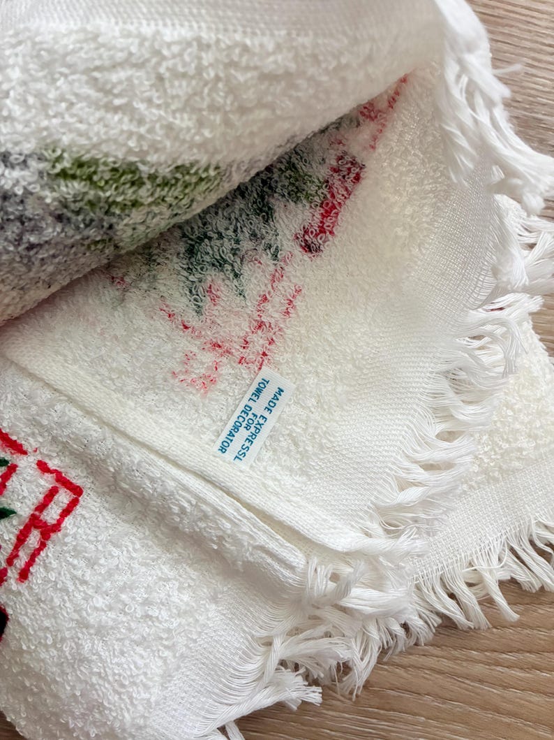 K&ouml;nnte beinhalten: Wei&szlig;es dekoratives Handtuch mit roten und gr&uuml;nen Stickereidetails. Das Handtuch hat einen Fransenrand und den Text "MADE FOR EXPRESSL TOWEL DECORATOR". Das Handtuch ist gefaltet und zeigt das gestickte Design.
