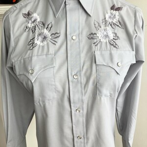 May include: A light blue long-sleeved button-down shirt with embroidered white flowers and grey leaves on the front. The shirt has a pointed collar and two chest pockets.