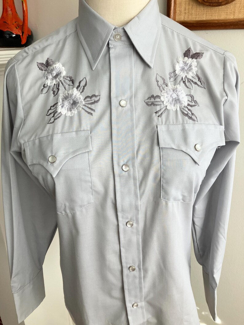 May include: A light blue long-sleeved button-down shirt with embroidered white flowers and grey leaves on the front. The shirt has a pointed collar and two chest pockets.