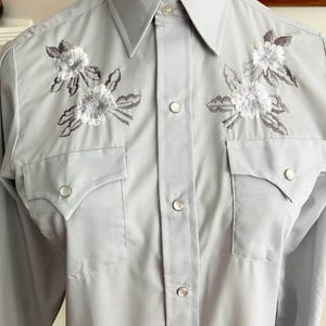 May include: A light grey long-sleeved button-down shirt with embroidered white flowers on the front. The shirt has a pointed collar and two chest pockets with button closures.