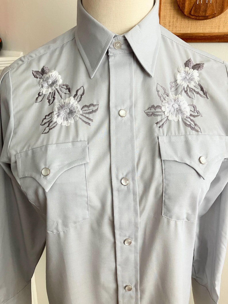 May include: A light grey long-sleeved button-down shirt with embroidered white flowers on the front. The shirt has a pointed collar and two chest pockets with button closures.