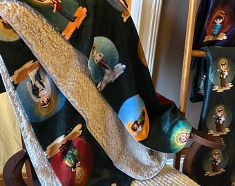 May include: A dark green blanket featuring a repeating pattern of saint illustrations within colourful circles. The blanket has a soft, light-coloured sherpa lining and the words "Saintly Snuggles" are printed on the blanket.