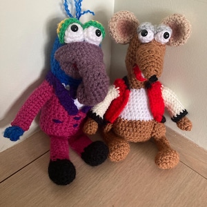 May include: Two handmade crochet dolls. The left doll has a purple nose, blue hair, and a magenta suit. The right doll is a brown mouse with large ears, wearing a white shirt and a red vest. Both dolls feature large white eyes with black pupils.