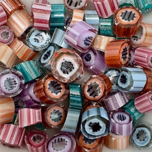 May include: A collection of colorful glass beads in various shapes and sizes. The beads feature stripes and swirls in shades of orange, pink, teal, purple, and blue. Some beads have a central design, while others are striped. The beads are likely for jewelry making.