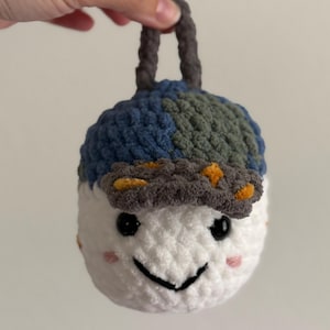 May include: A handmade, white crocheted snowman ornament with a blue, green, and gray hat. The snowman has a smiling face with black eyes and a black mouth. The ornament has a gray loop for hanging.