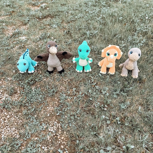 May include: A set of five crocheted dinosaur toys in various colors including bright blue, brown, light green, orange, and beige. Each stuffed animal features dark safety eyes and distinct shapes representing different prehistoric creatures.