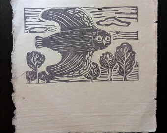 May include: A square piece of off-white paper with a deckled edge featuring a dark gray linocut print. The artwork depicts a stylized owl in flight above a landscape of trees, with horizontal lines in the background representing the sky.