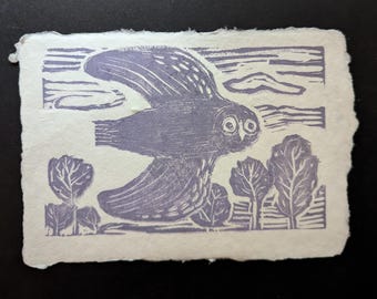 May include: A rectangular print on off-white textured paper featuring a purple ink illustration of an owl in flight. The owl is depicted above stylized trees and clouds, with visible carved linoleum texture throughout the purple ink design.