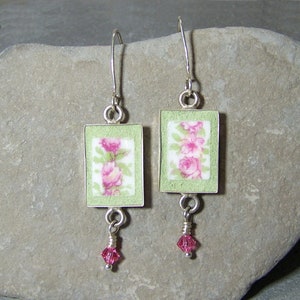 May include: A pair of silver-toned earrings. Each earring features a rectangular, light green frame with a floral design in pink and green. A small pink crystal dangles from each earring. The earrings are displayed on a gray stone surface.