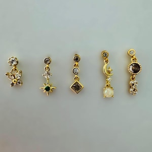 Könnte beinhalten: Fünf goldfarbene Ohrringe mit verschiedenen Designs. Die Ohrringe verfügen über klare und farbige Edelsteine, darunter eine Blume, ein Stern und ein Mond. Die Ohrringe sind für durchbohrte Ohren konzipiert und werden vor einem weißen Hintergrund präsentiert.