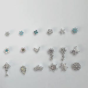 May include: A collection of silver-toned stud earrings in various floral and geometric designs. Some earrings feature clear and turquoise-colored gemstones. The earrings are arranged on a white surface, showcasing their intricate details and sparkling appearance.