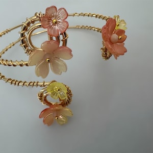 May include: A gold-toned arm cuff with a spiral design, adorned with translucent, flower-shaped accents in shades of peach and yellow. The floral elements are clustered around the cuff's loops, creating a delicate and feminine aesthetic.