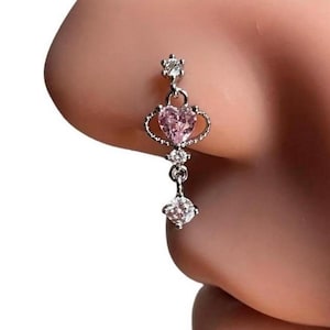 Puede incluir: Un aro de nariz plateado con una piedra rosa en forma de corazón y detalles de cristal transparente. El aro tiene un dije colgante con una piedra de cristal transparente.
